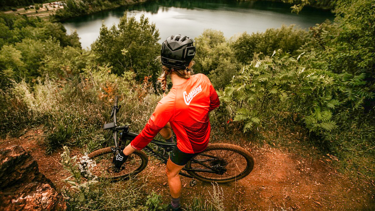 Midwest Mountain Bike Fest-Scenic Overlook A cyclist in a red jersey and helmet sits on a mountain bike at the edge of the Cuyuna mountain bike trails, overlooking a lush forest and lake below—a scenic view from behind, perfect for the Midwest Mountain Bike Fest.