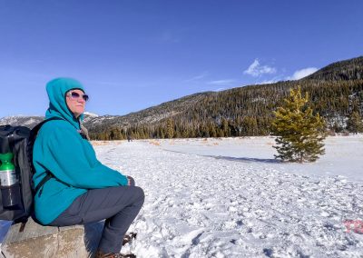 A person in a teal jacket, sunglasses, and Breakwater Supply Fogland Backpack sits on a rock in a snowy landscape, surrounded by mountains and pine trees under a clear blue sky.