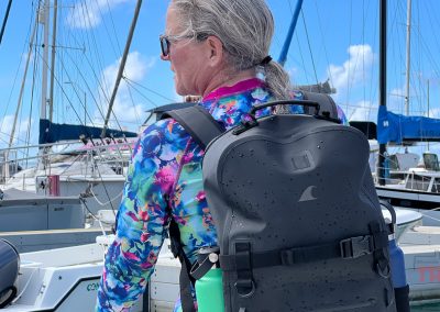 A person with gray hair in a ponytail, wearing sunglasses and a colorful floral shirt, stands on a marina dock with a black Breakwater Supply Fogland Backpack. Sailboats and blue sky are visible in the background.