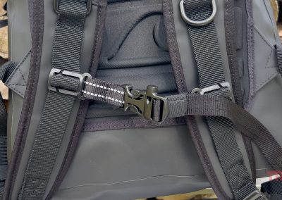 Close-up of the Breakwater Supply Fogland Backpack's padded shoulder straps and chest buckle, set against a background of stacked logs. The backpack features visible stitching and hardware details.