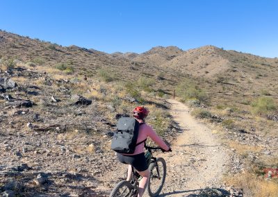 A person wearing a red helmet and Breakwater Supply Fogland Backpack rides a mountain bike on a rocky dirt trail through a dry, hilly desert landscape under a clear blue sky.