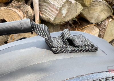 Close-up of a black, textured strap attached to the matte black Breakwater Supply Fogland Backpack, with stacked firewood in the blurry background.
