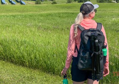 A woman with a ponytail, wearing a hat and pink long-sleeve shirt, stands in a grassy field. She carries a large black Breakwater Supply Fogland Backpack with a tripod attached and holds a green water bottle. Blue structures are visible in the background.