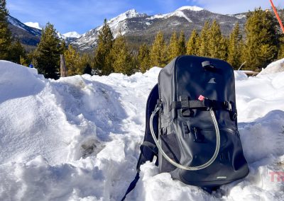 A black Breakwater Supply Fogland Backpack with a hydration tube sits on snow, surrounded by pine trees and snowy mountains under a blue sky.