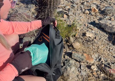 A person in a pink long-sleeve shirt and gloves sits on rocky ground, opening a Breakwater Supply Fogland Backpack filled with hiking gear, including what appears to be a green jacket. Desert plants and rocks surround them.