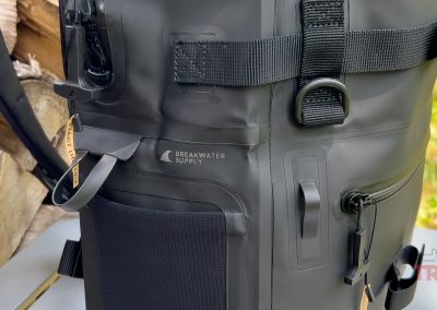 Close-up of a black Breakwater Supply Fogland Backpack showing straps, buckles, and attachment points, with part of a wooden log in the background.