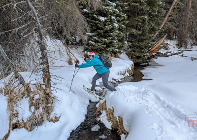 A person in winter gear with a Breakwater Supply Fogland Backpack uses ski poles to cross a narrow, snow-covered creek in a snowy forest. Another person stands nearby.