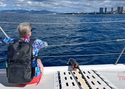 A person with gray hair, wearing a colorful shirt and a Breakwater Supply Fogland Backpack, sits on the deck of a boat looking out at the ocean, with a city skyline and mountains visible in the distance under a partly cloudy sky.
