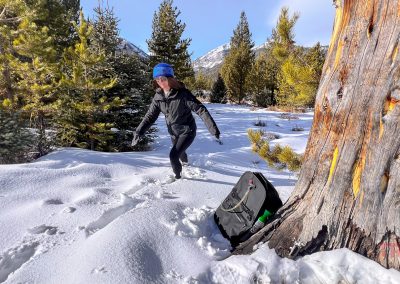 A person wearing winter clothing and a blue hat walks through deep snow in a pine forest with mountains beyond; a Breakwater Supply Fogland Backpack rests against a tree in the foreground.