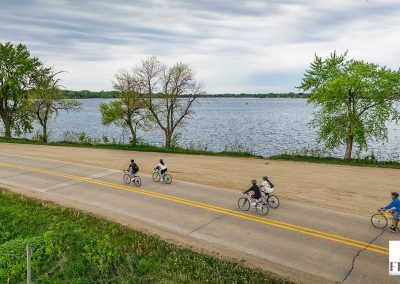 Five people enjoy the Prairie Pedal Bike Ride along a paved road beside a large lake bordered by trees under a cloudy sky.