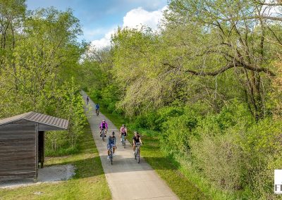 A group of cyclists enjoy the Prairie Pedal Bike Ride along a paved trail surrounded by lush green trees on a sunny day. A small wooden shelter sits beside the path.
