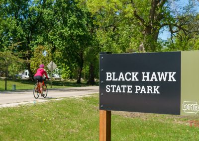A cyclist wearing a pink jacket rides on a paved path near a large sign that reads Black Hawk State Park DNR in Sac County, Iowa, surrounded by green trees and grass on a sunny day.