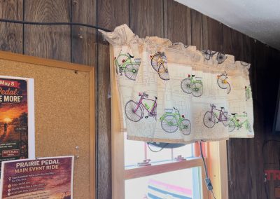 A wood-paneled room at Trailside Roadhouse in Carnarvon, IA, with a window draped in a curtain of colorful bicycle designs. A corkboard displays Prairie Pedal posters, and part of a TV screen showing a person is visible.