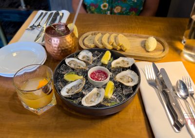 A restaurant table at Cattlemen's Steak House in Sac City, IA is set with oysters on ice, lemon wedges, sauce, sliced bread with butter, a cocktail in a copper mug, an orange-garnished drink, and cutlery; a person in a floral dress sits behind.