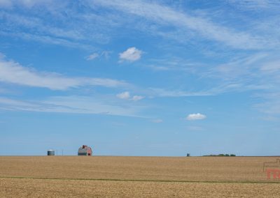 A wide, flat field of harvested crops under a blue sky with scattered clouds. In the distance, a red barn and silos near the horizon hint at rural life along the Prairie Pedal Bike Ride in Sac County, Iowa.