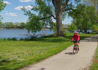 A person wearing a pink top and helmet rides a bike on a paved riverside trail by Black Hawk Lake in Lake View, IA, surrounded by green grass, trees, and a blue sky with clouds.