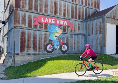 A cyclist in a pink outfit rides past a large metal building with a mural of a blue bird riding a bicycle and a sign that reads Lake View IA on a sunny day.