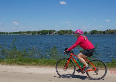 A woman wearing a pink helmet and sweatshirt rides an orange bicycle along a paved path beside Black Hawk Lake in Lake View, IA, with trees and houses in the background under a clear, sunny sky offering a beautiful lake view.