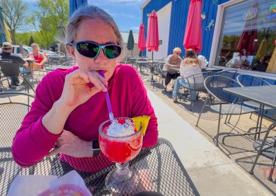 At Cielito Lindo Restaurant in Lake View, IA, a person wearing sunglasses and a pink top sips a bright red frozen drink with whipped cream and fruit garnish at an outdoor café with metal tables and umbrellas on a sunny day.