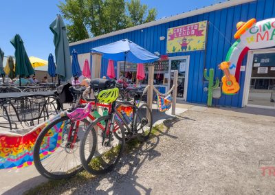 Bicycles are parked outside Cielito Lindo Restaurant in Lake View IA, a blue eatery decorated with colorful banners and a Cinco de Mayo arch. People are sitting at tables under umbrellas on a sunny day.