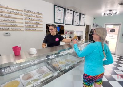 At Houdini's Sandwich Shop in Lake View, IA, a woman in colorful shorts receives an ice cream cone from a smiling employee behind the counter at a bright, modern ice cream shop with a menu and tubs of ice cream visible.