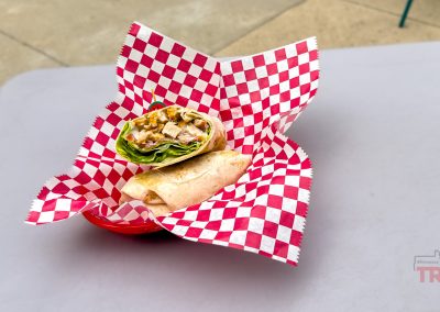 A wrap sandwich cut in half sits in a red basket lined with red-and-white checkered paper at Houdini's Sandwich Shop, placed on a light gray table in Lake View, IA.