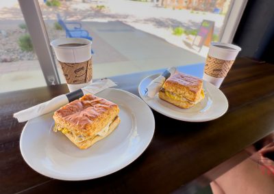 Two plates with breakfast sandwiches and two cups of coffee from Pura's Brewtique sit on a wooden counter by a window in Lake View Ia, sunlight streaming in as napkins and utensils rest on the plates.