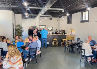 A busy café, Pura's Brewtique in Lake View IA, with people sitting at tables, eating and talking. Some stand in line near the counter at the back. The space has high ceilings, white walls, and large windows flooding it with natural light.