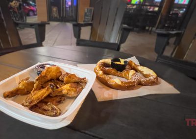 A tray of chicken wings with dipping sauce and a pretzel with cheese sauce sit on a dark outdoor table at Soak'd Bar and Grill, Lake View IA, with wooden chairs on the restaurant patio at night.