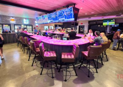 A lively bar in Lake View IA, Soak'd Bar and Grill features a glowing pink countertop, patrons on padded barstools, TVs displaying sports, and groups chatting and dining under warm lighting.