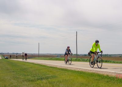 Cyclists ride along a rural road in Sac County, Iowa, during the Prairie Pedal Bike Ride. Green fields line both sides under a partly cloudy sky as a person in a bright yellow jacket leads the group, with more cyclists and distant cars in view.