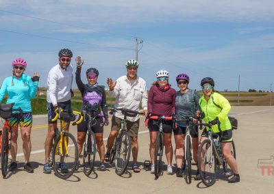 A group of seven cyclists, wearing helmets and colorful outfits, stand with their bikes on a rural roadside in Sac County Iowa, smiling and waving at the camera under a blue sky during the Prairie Pedal Bike Ride.