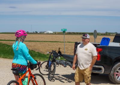 A woman in a bright helmet and colorful shorts stands with her bike, chatting with a man by a parked pickup truck on a rural road near farm fields during the Prairie Pedal Bike Ride in Sac County, Iowa.