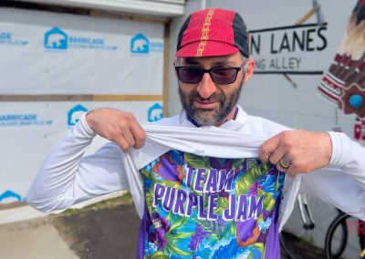 A man wearing sunglasses and a red cycling cap lifts his white jacket to reveal a colorful “Team Purple Jam” shirt. He stands outside next to mural-covered walls, ready for the Prairie Pedal Bike Ride in Sac County, Iowa.