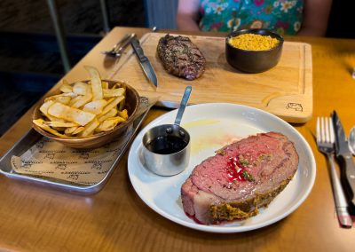 At Cattlemen's Steak House in Sac City, IA, a table is set with rare roast beef and sauce, thick-cut fries, and a wooden board holding grilled steak and macaroni and cheese. A person sits behind the feast.