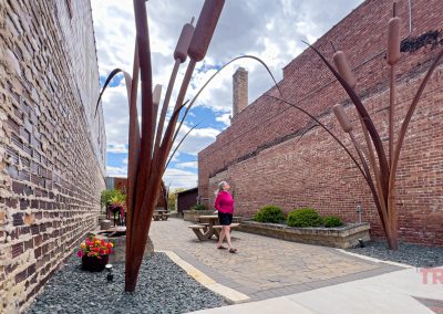 A person stands in a narrow, outdoor space between brick buildings in Sac City, Sac County Iowa, surrounded by tall metal sculptures resembling cattails. Benches, potted flowers, and a clear sky are visible.