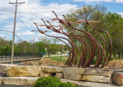 A metal sculpture of abstract, flowing shapes stands on a stone platform near a sidewalk and bridge in Sac City, Sac County Iowa, surrounded by grass, trees, and utility poles under a partly cloudy sky.