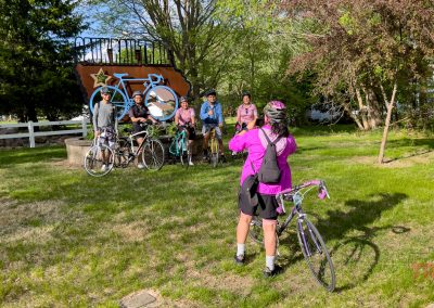 A group of five cyclists pose with their bikes by a large bicycle sculpture on the Sauk Rail Trail, while a sixth cyclist in a pink jacket snaps their photo during the sunny Prairie Pedal Bike Ride in a green park.