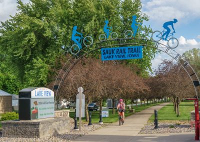 A cyclist rides under a decorative arch reading Sauk Rail Trail, Lake View, Iowa with blue bike figures on top. Trees and signs line the paved trail on a sunny day during the Prairie Pedal Bike Ride.