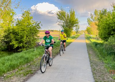 Two people wearing helmets ride bicycles on the Sauk Rail Trail through a green, tree-lined landscape under a partly cloudy sky. The bright, inviting scene suggests a perfect day for outdoor activity during the Prairie Pedal Bike Ride.