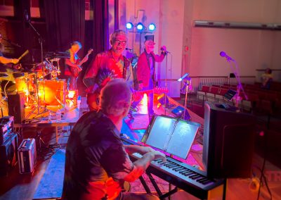 A band performs on stage with colorful lights at the Shull Center in Sac City, IA. The keyboardist plays in the foreground, while a singer, guitarist, and drummer are visible. Some empty seats can be seen in the audience area.