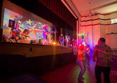 A band performs on stage at the Shull Center in Sac City, IA under colorful lights, as a few people dance in front. The theater's red and cream walls, empty seats, and a star-studded mural with musical motifs complete the lively scene.