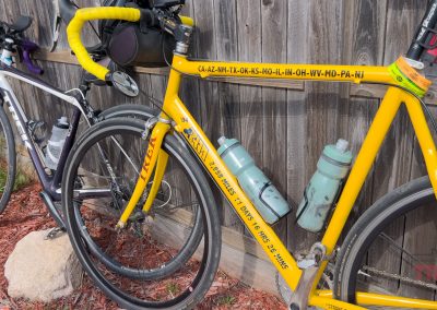 Two bicycles are parked against a wooden fence, ready for the Prairie Pedal Bike Ride. The closer bike is bright yellow with text on the frame and two water bottles; the other is gray with blue accents and also has a water bottle attached.