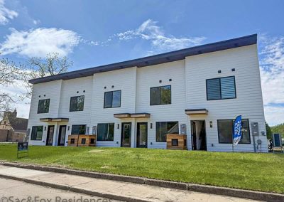 Modern two-story white townhouse building with large windows, four entrances, a small lawn in front, and a blue “open house” flag. Sac and Fox Residences in Sac City IA stand out against a clear sky with some clouds in the background.