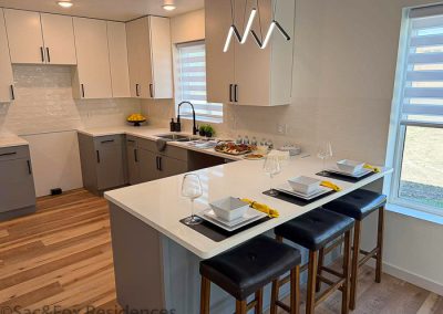 Modern model kitchen with white cabinets, light wood flooring, and a large island set for three with white dishes and wine glasses. Pendant lights hang above the island, and sunlight streams in through window shades at Sac and Fox Residences in Sac City, IA.