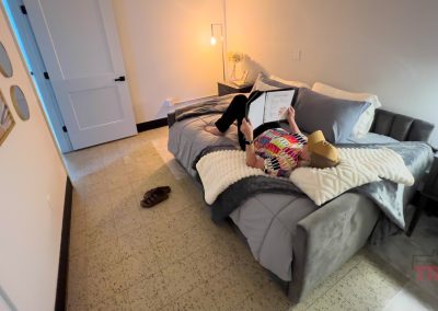 A woman lies on a neatly made bed in a cozy Sac and Fox Flats bedroom, reading a book while holding a stuffed animal. A lamp glows softly on the nightstand, slippers rest nearby, and the clock reads 4:37.