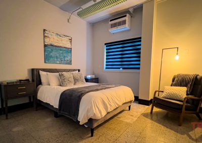 A modern bedroom at Sac and Fox Flats features a bed with a gray headboard, white and gray bedding, a nightstand with books, a cozy chair with pillow and blanket, small lamp, painting above the bed, and a window with black blinds.