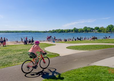A person wearing a pink shirt and helmet rides a bicycle on a paved path near Lakeville MN's Antlers Park, while people relax, walk, and swim by the water under a clear blue sky. Trees and houses line the opposite shore.