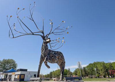 A large metal sculpture of a reindeer with elaborate, branching antlers stands outdoors in Antlers Park, Lakeville MN, with a blue sky and several buildings and trees in the background.