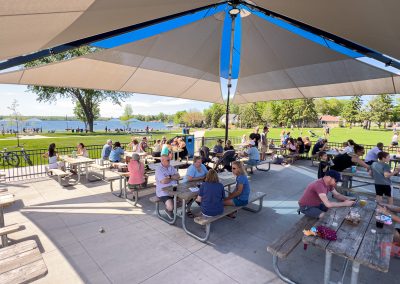 People sit at picnic tables under a large pavilion in Antlers Park, Lakeville MN, enjoying food and drinks. The park is busy with others walking, playing, and relaxing near a lake on a sunny day, surrounded by trees and green grass.